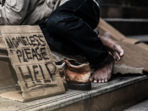 A man lies on some steps next to a sign saying "Homeless - please help"