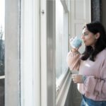 woman looks out of window with mug in her hand