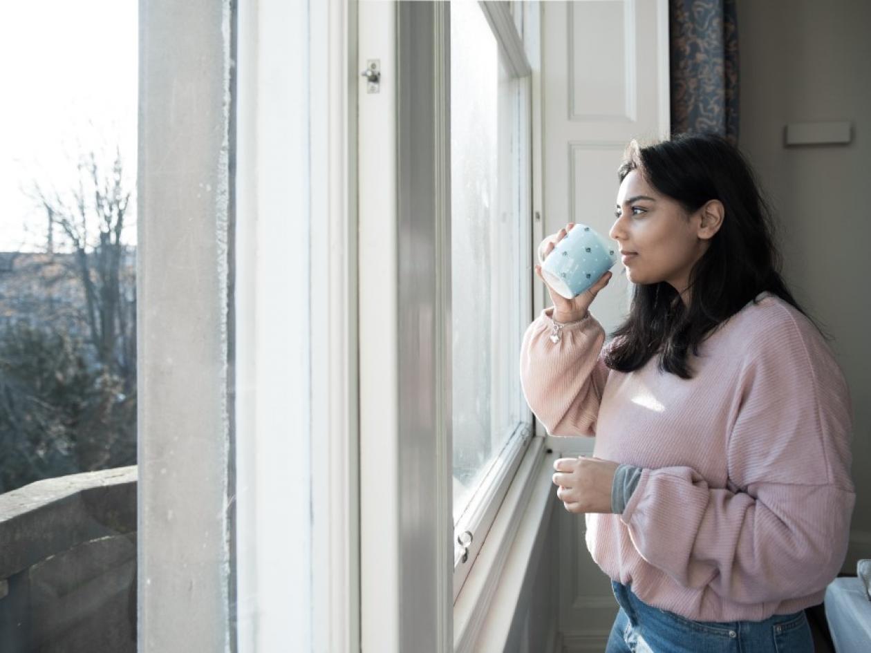 woman looks out of window with mug in her hand