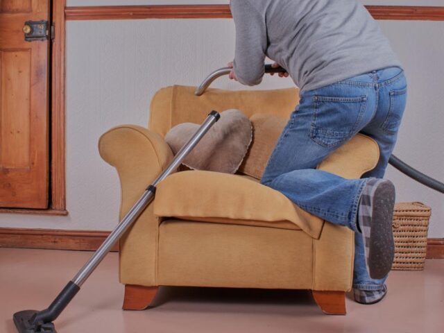 a man kneels on an armchair as he hoovers its upholstery