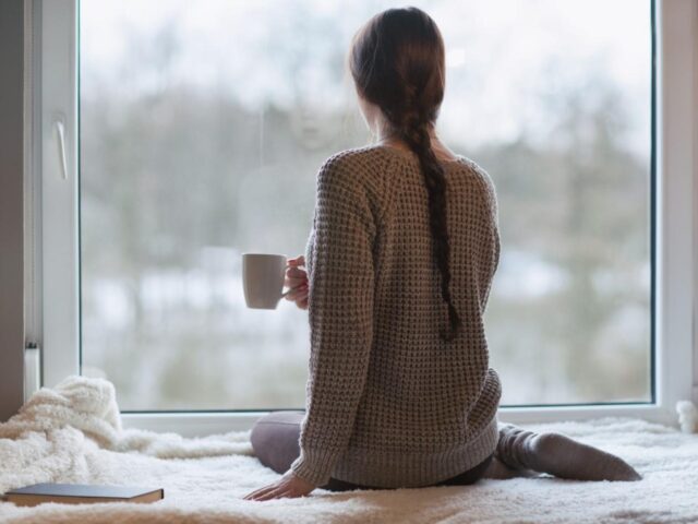 a woman sits on a bed holding a mug and looking out of a window with her back to us