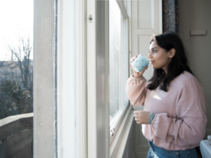A woman looks out of a window while drinking from a mug