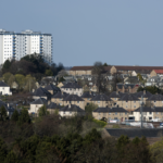 A view across a town with tower blocks in the background and houses in the foreground. There are trees among the buildings, and a blue sky