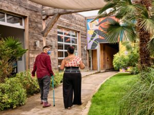 Two masked disabled people of color chat and walk through an outdoor courtyard, photographed from the back. One walks with a cane.