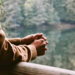 someone leans on a balcony overlooking a lake with trees around it. We see only their arms. Photo by Ümit Bulut on Unsplash