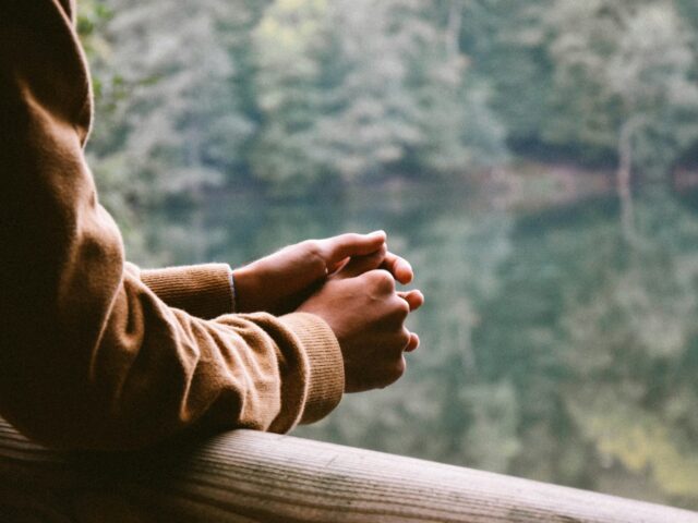 someone leans on a balcony overlooking a lake with trees around it. We see only their arms. Photo by Ümit Bulut on Unsplash