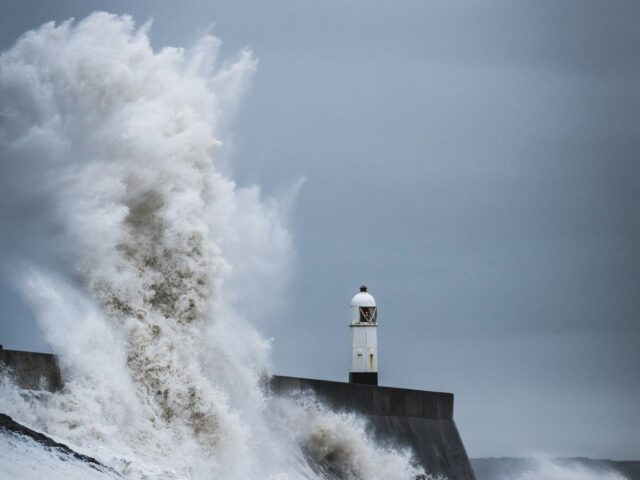 a storm wave crashes on a seafront with a lighthouse in the background